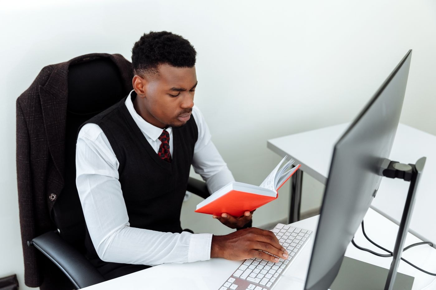 A person reading a book beside a computer